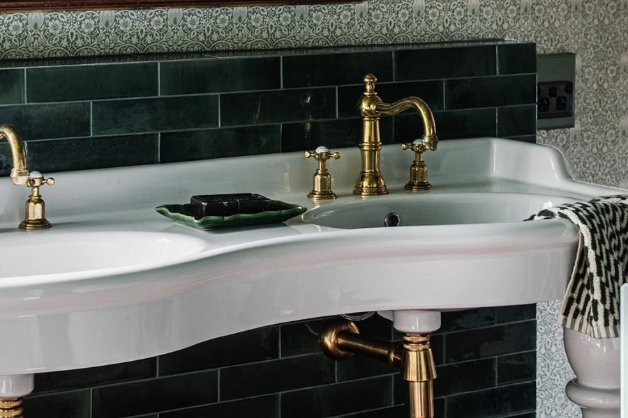 Golden brass basin taps by Perrin and Rowe catch the eye against a curved white twin basin vanity in green tiled heritage bathroom.
