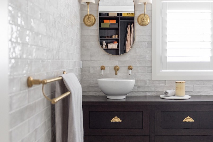 Wall mounted natural brass basin taps fill vessel bowl basins on dark wooden vanity alongside double towel rail made from brass.
