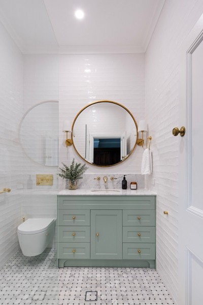 Polished brass art deco style basin taps are wall mounted above marble basin counter with pale green cabinets.
